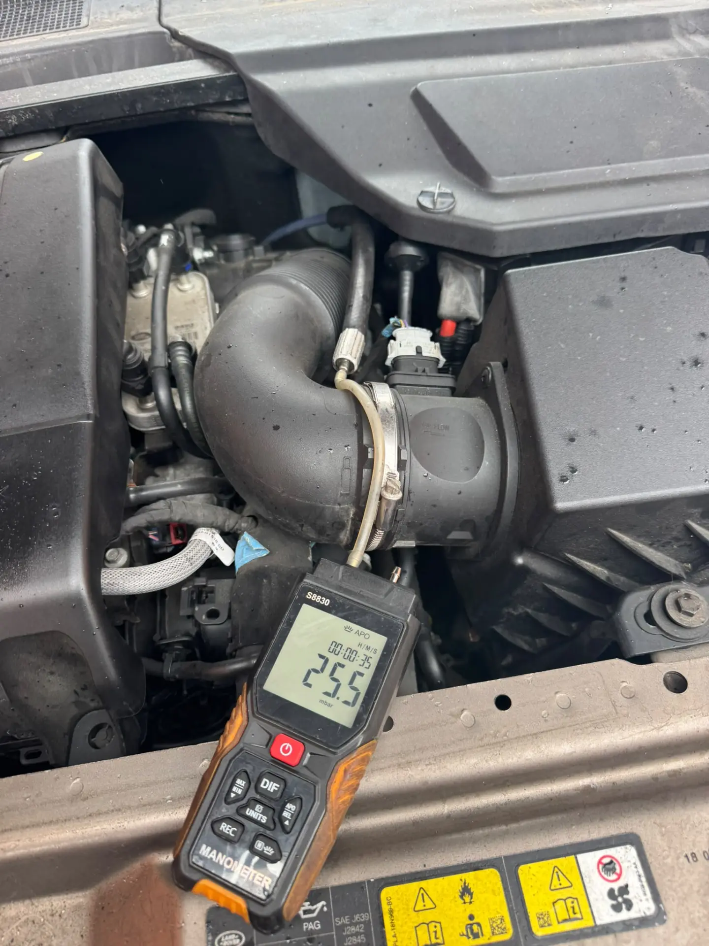 Engine bay of a Land Rover Discovery Sport during a DPF diagnostic inspection, showing a digital manometer connected to the system with a pre-cleaning pressure reading of 25.5 mbar.
