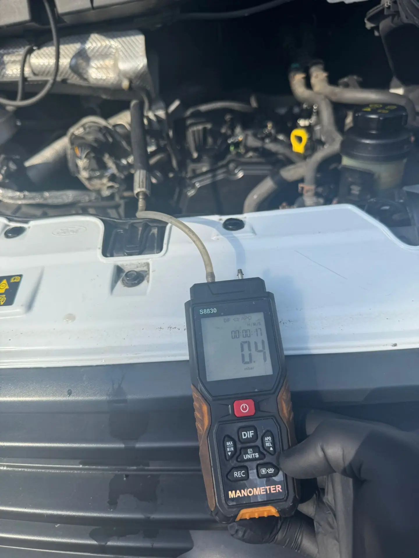Technician performing DPF pressure testing on a Ford Transit engine bay using a digital manometer, showing a post-cleaning reading of 0.4 mbar.
