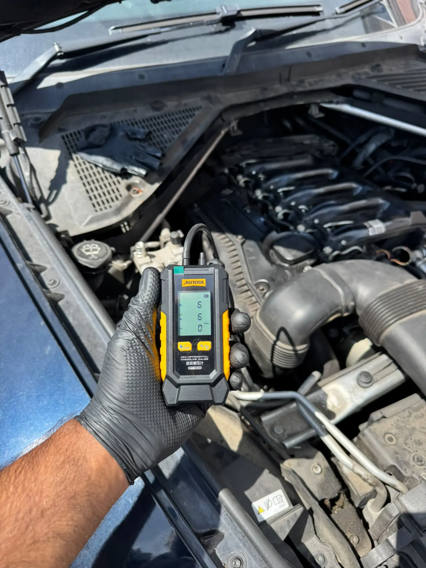 Technician holding a digital DPF pressure gauge while testing the engine of a BMW X5 before DPF cleaning.